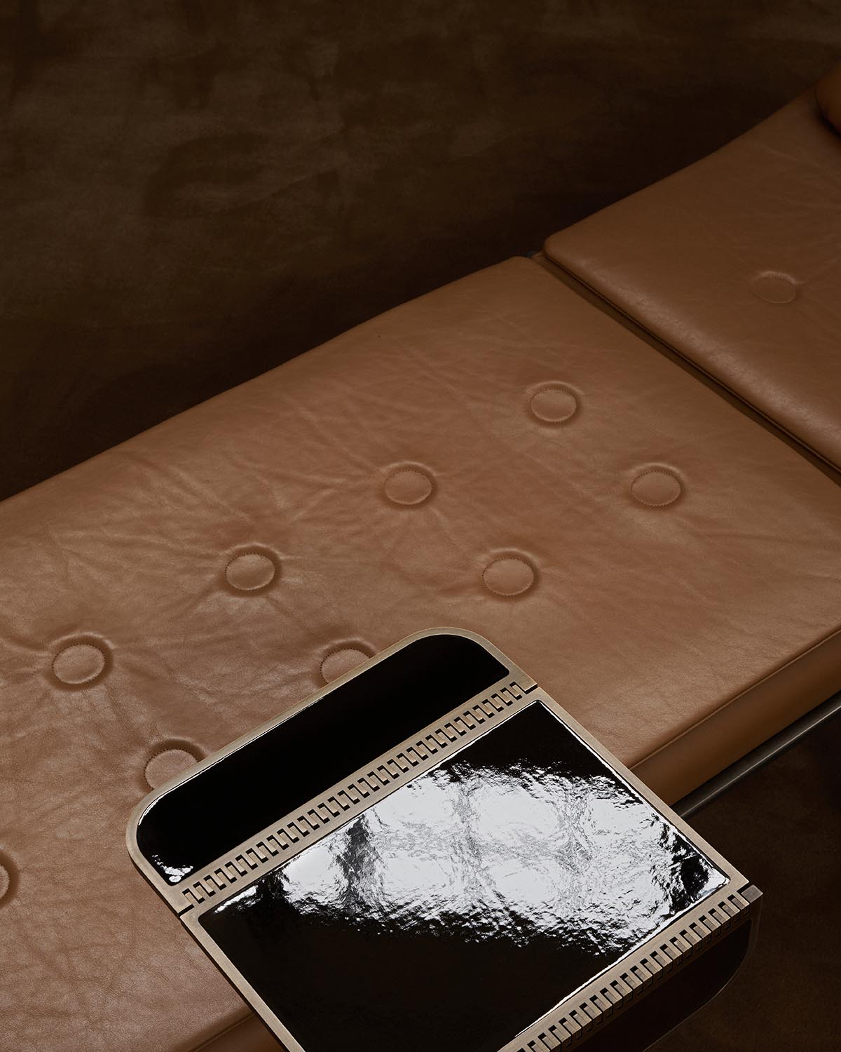 Close-up of a brown tufted leather bench with a glossy black rectangular tray attached on one side against a dark background