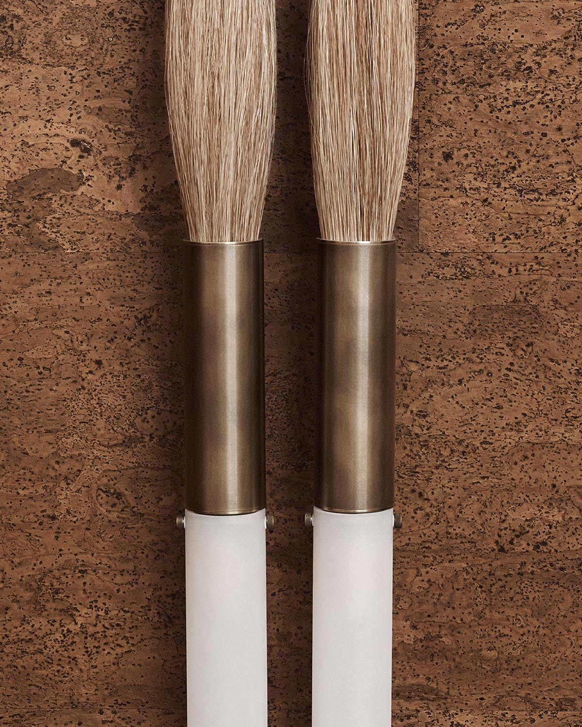 Close-up of two natural bristle brushes with bronze metal ferrules and white handles against a textured brown background