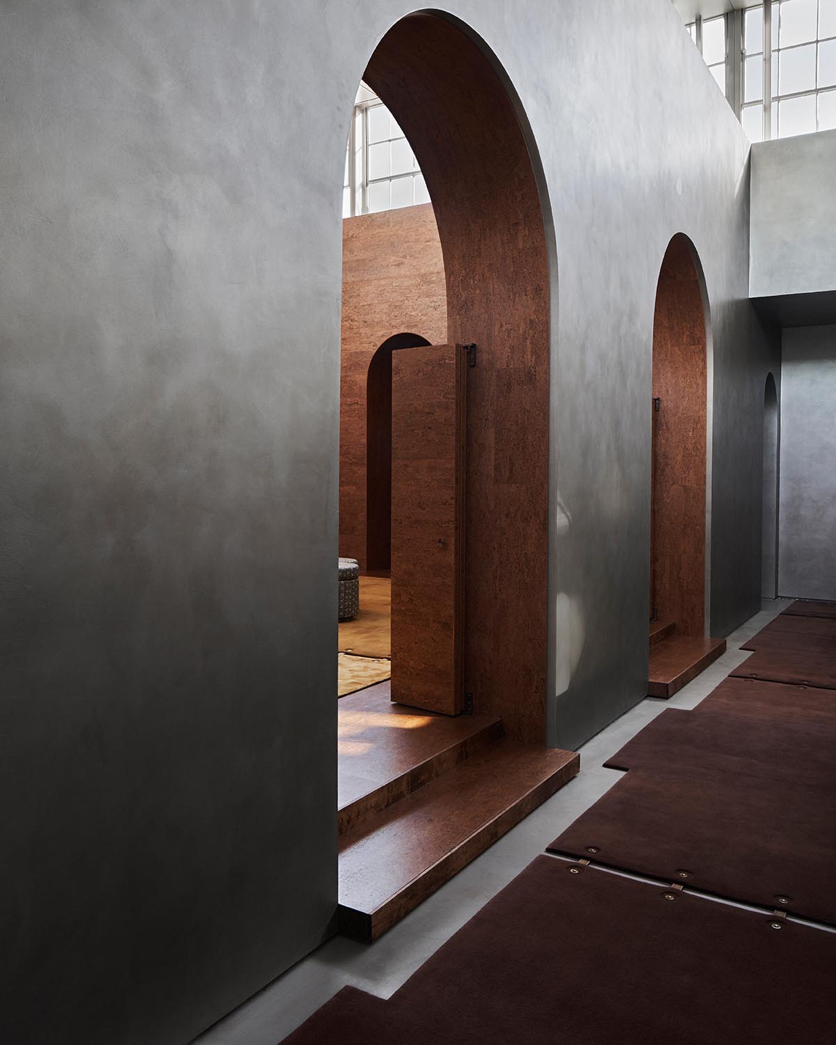 Interior view of a modern hallway with grey plaster walls, large arched wooden doorways, and natural light from high windows