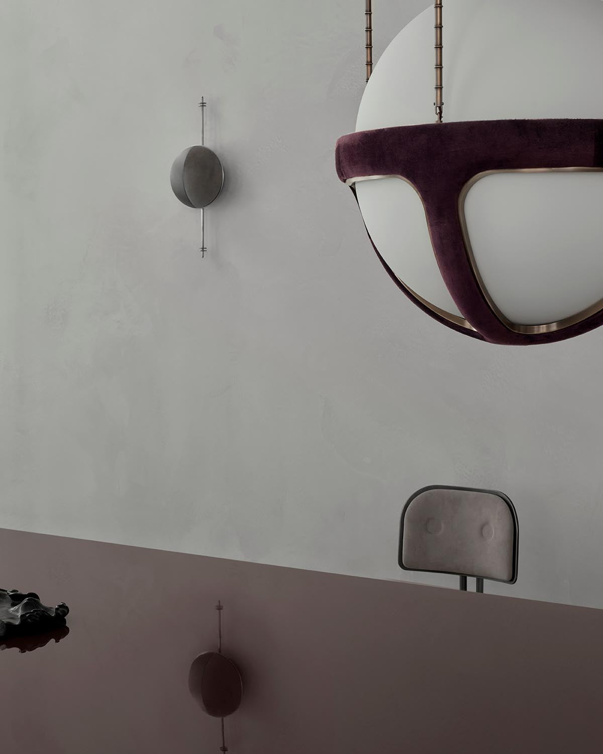 Minimalist interior showing a suspended white and burgundy pendant light, muted gray wall, and a partial chair backrest