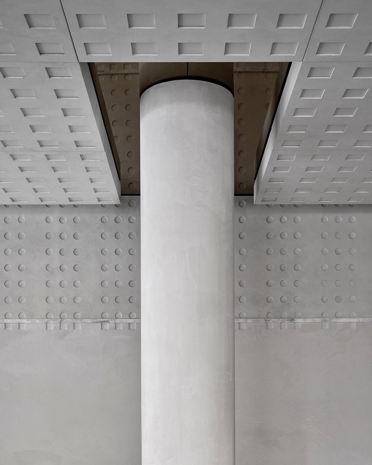 Close-up of a smooth cylindrical concrete pillar centered between textured walls and ceiling panels with geometric patterns