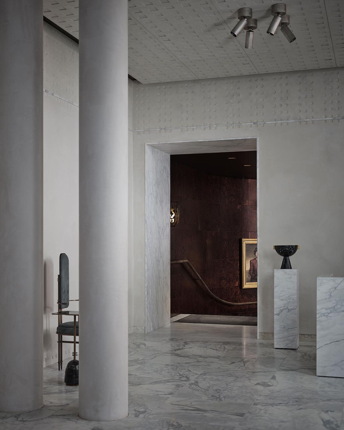 Interior view of a minimalist space with tall cylindrical columns, marble flooring, and a dark stairway entrance with framed artwork visible