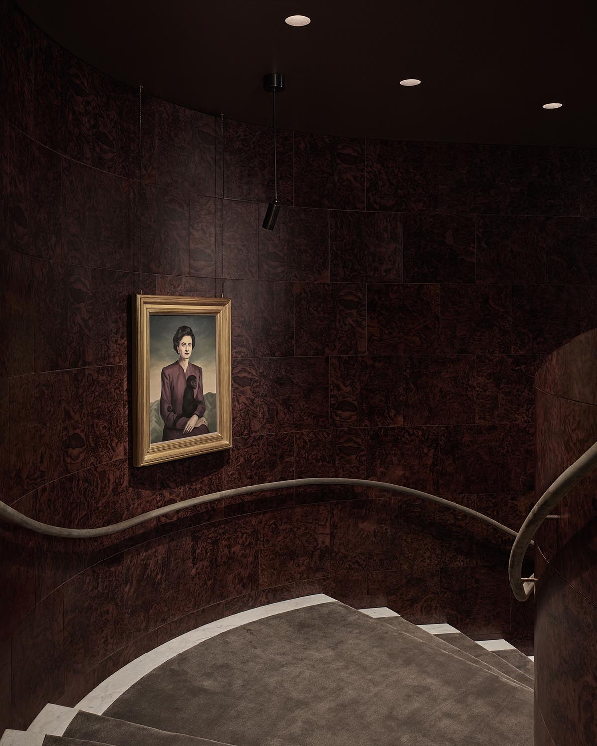 Curved staircase with carpeted steps and dark wood-paneled walls featuring a framed portrait under spotlight