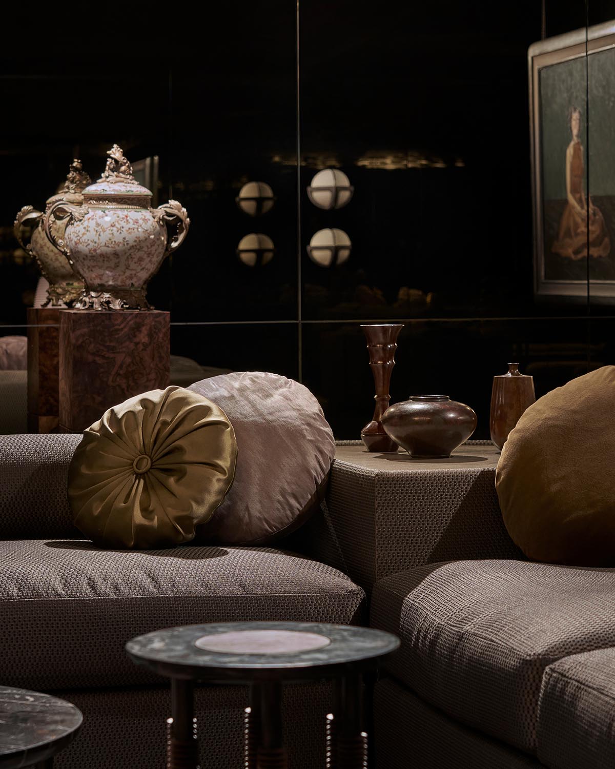 Cozy seating area with textured neutral sofas, round decorative pillows, ceramic vases, and a framed artwork on dark reflective wall