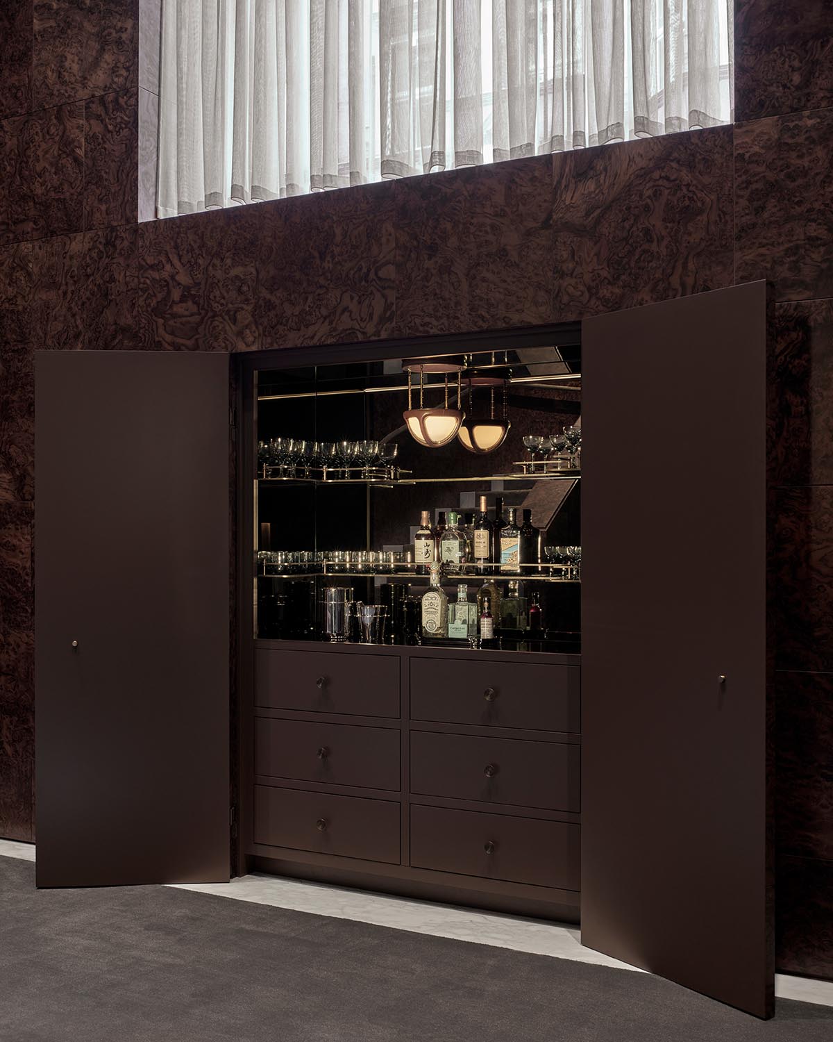 Dark built-in liquor cabinet with open double doors revealing bottles, glassware, and drawers against a textured wall