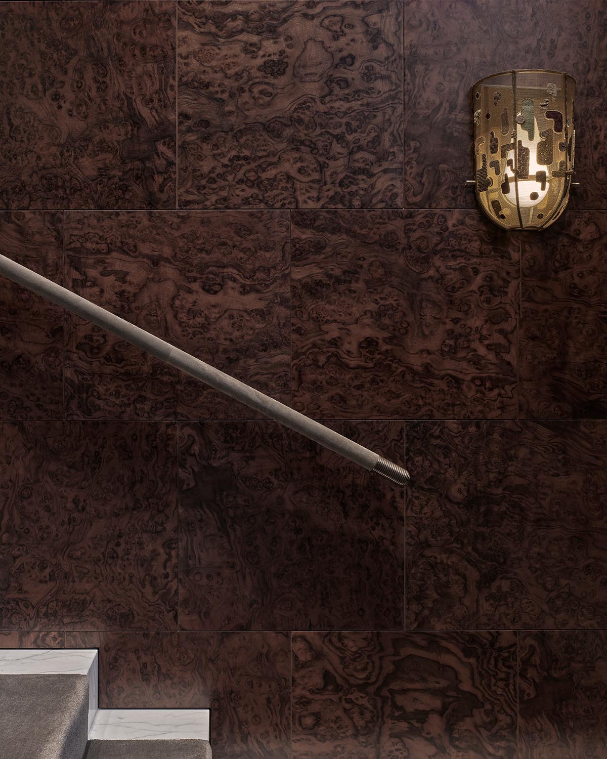 Dark textured wall panels with a metallic patterned wall sconce and a simple metal handrail over light stone stairs