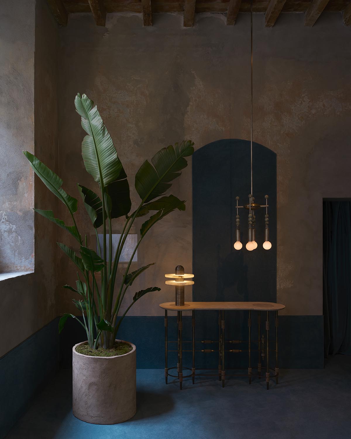 Indoor corner with large potted banana plant, wooden table with brass base, pendant light, and textured walls