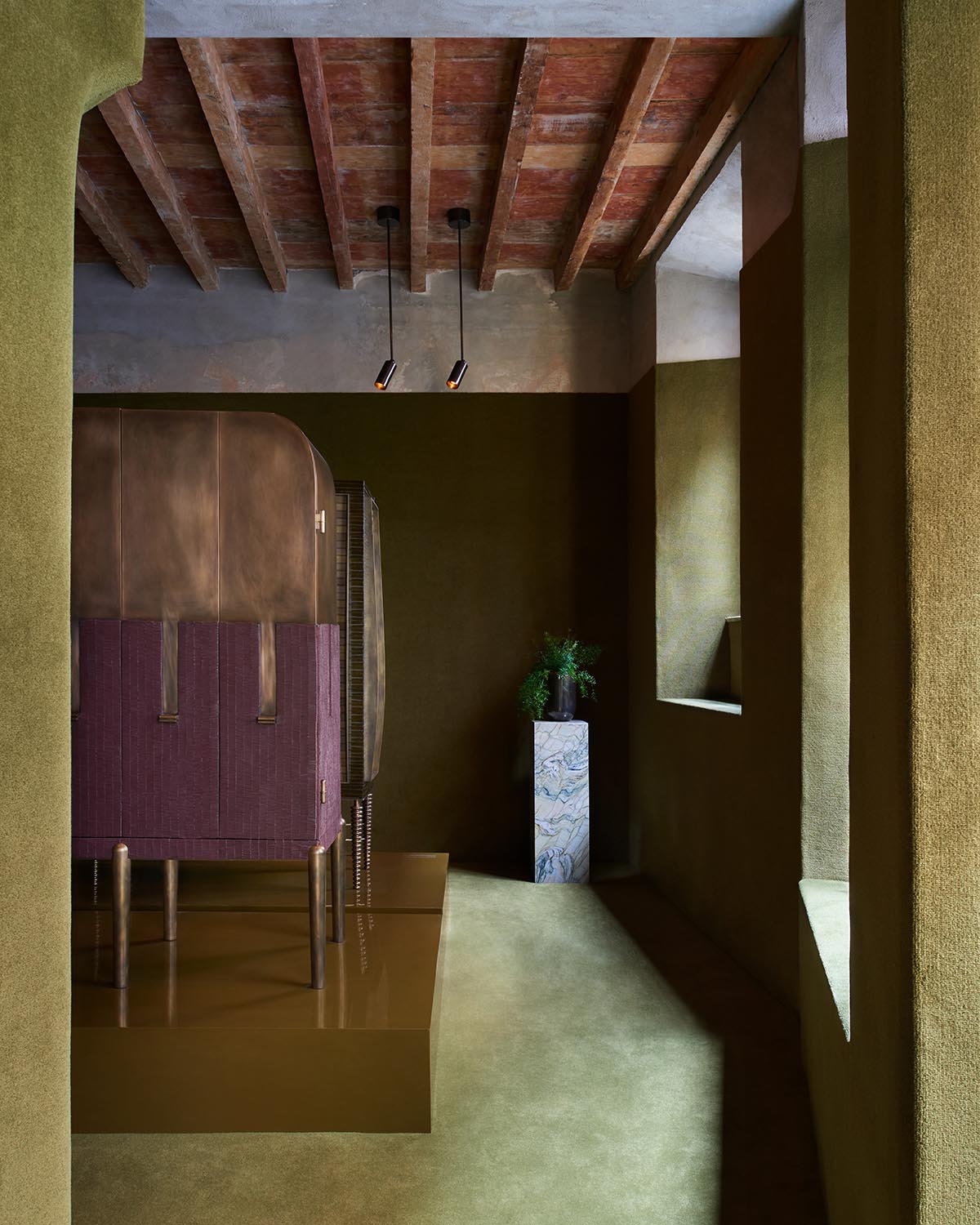 Interior view of a room with textured olive-green walls, wooden ceiling beams, and modern pendant lights over stylized furniture on a platform