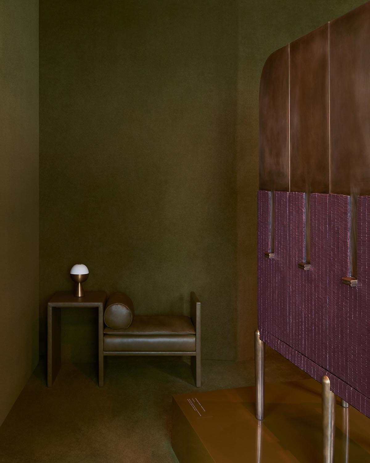 Minimalist corner with olive green walls, a brown leather bench, side table, white lamp, and a textured brown divider panel.