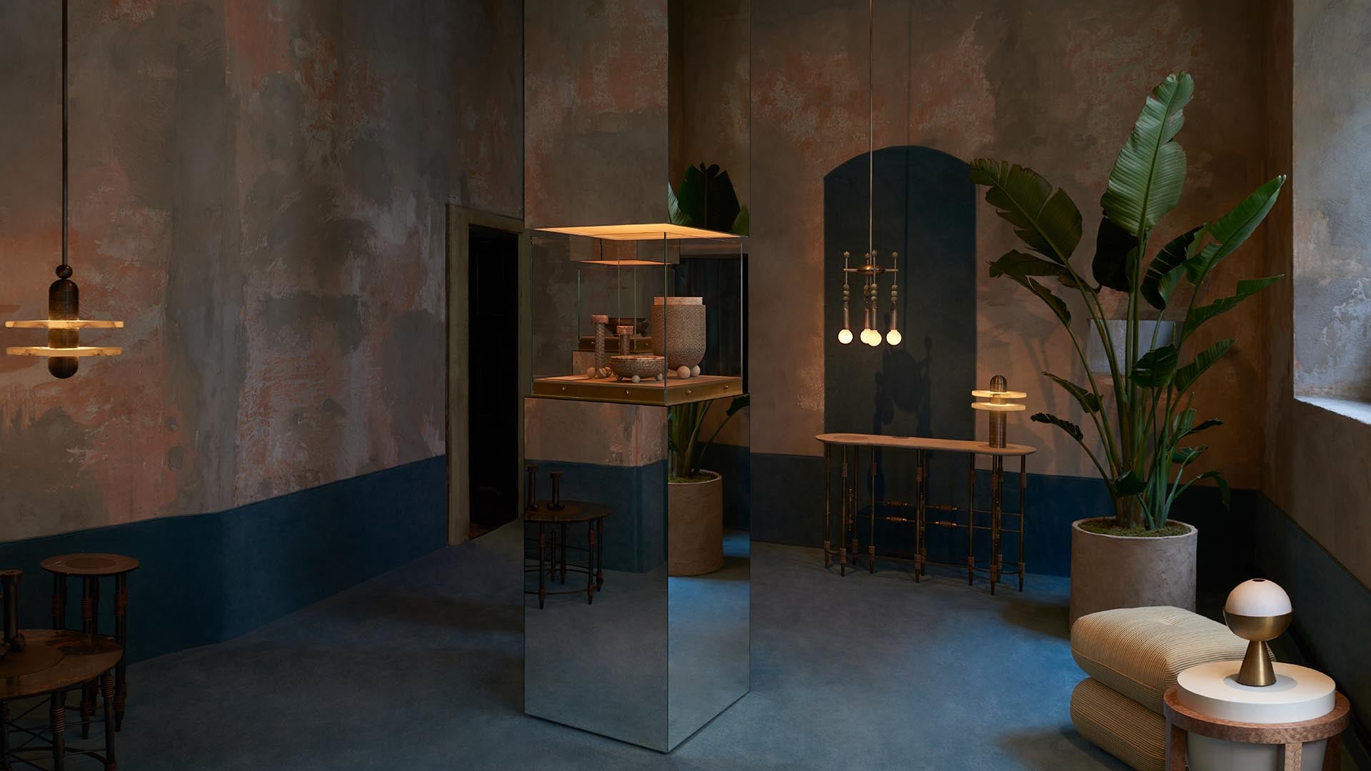 Dimly lit room with textured walls, large potted plants, mirrored display pedestal, and modern pendant lights hanging above wooden furniture