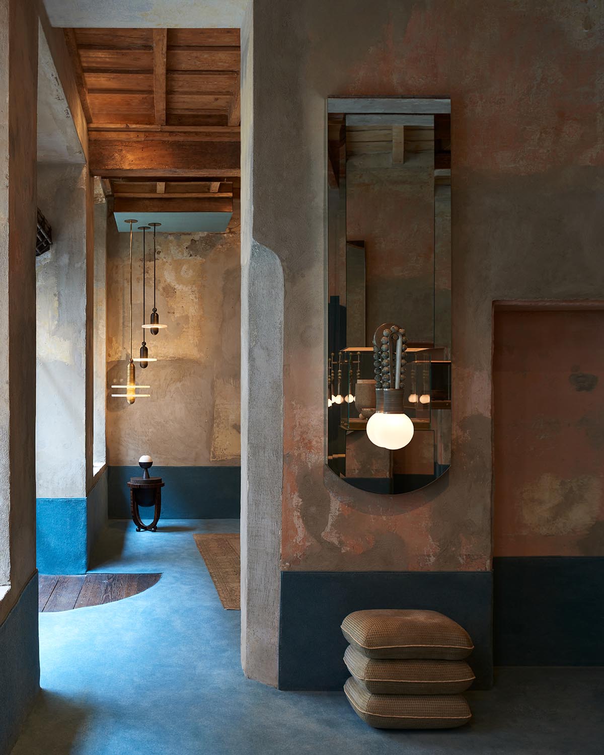 Interior space with textured walls, wooden ceiling beams, pendant lights, a tall mirror, and stacked cushions on a blue floor.
