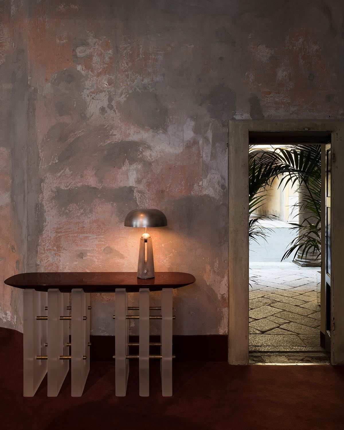 Sparse room with textured worn walls, featuring a sculptural table with a lit lamp beside an open doorway showing outdoor plants.