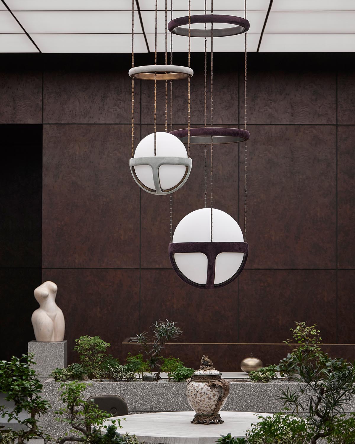 Contemporary pendant lights with spherical white shades suspended from circular frames above a stone planter with greenery and sculpture