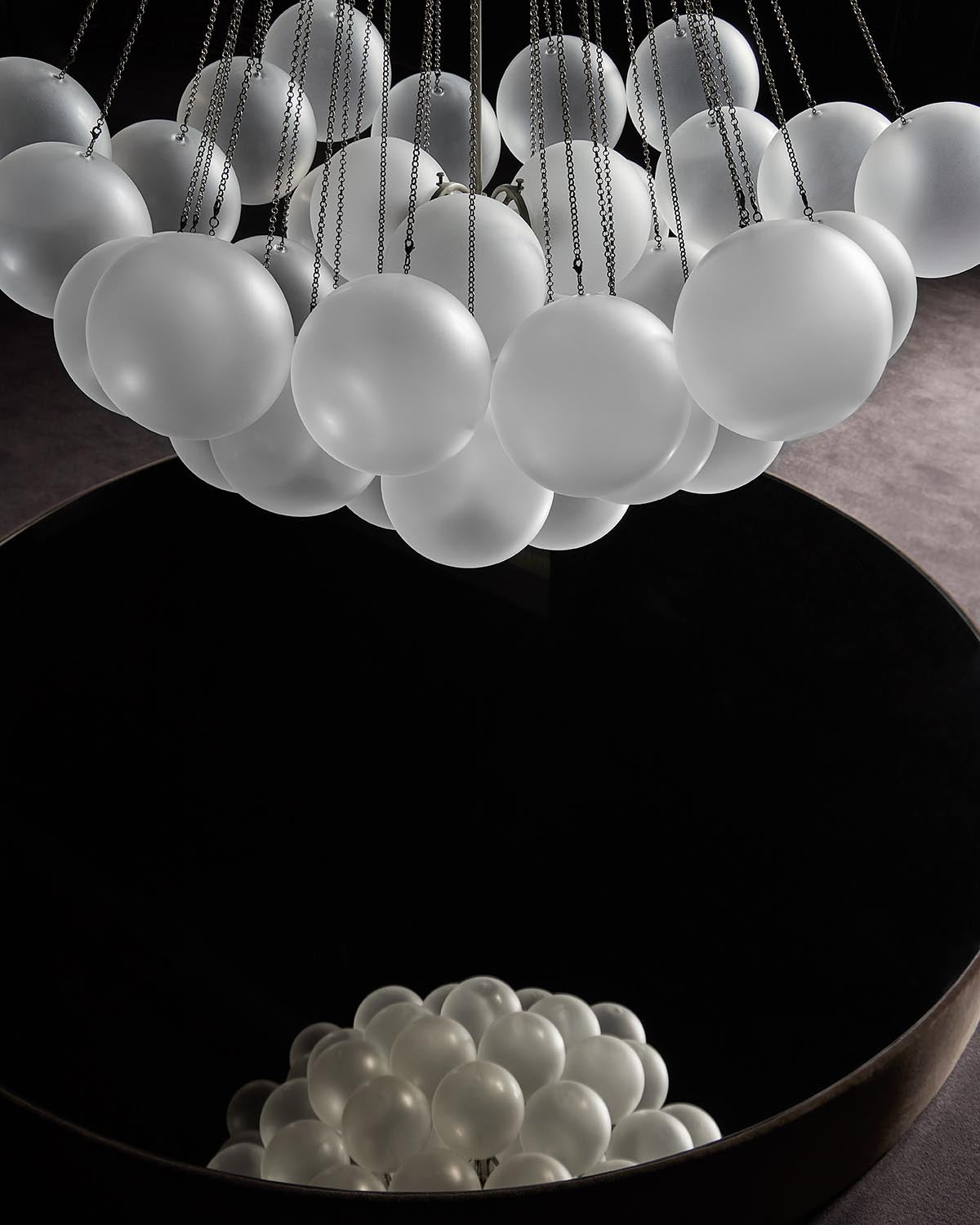 Cluster of frosted glass spheres suspended by metal chains above a large circular reflective surface in a dark space