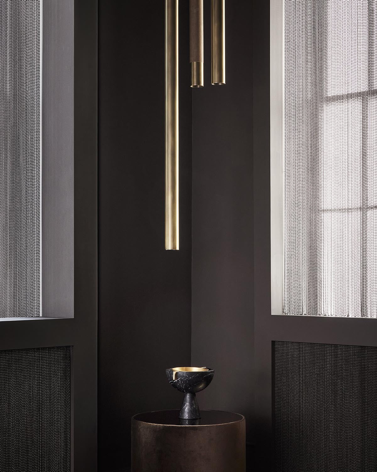 Black marble chalice on a dark cylindrical pedestal beneath three suspended brass cylindrical lights against dark walls