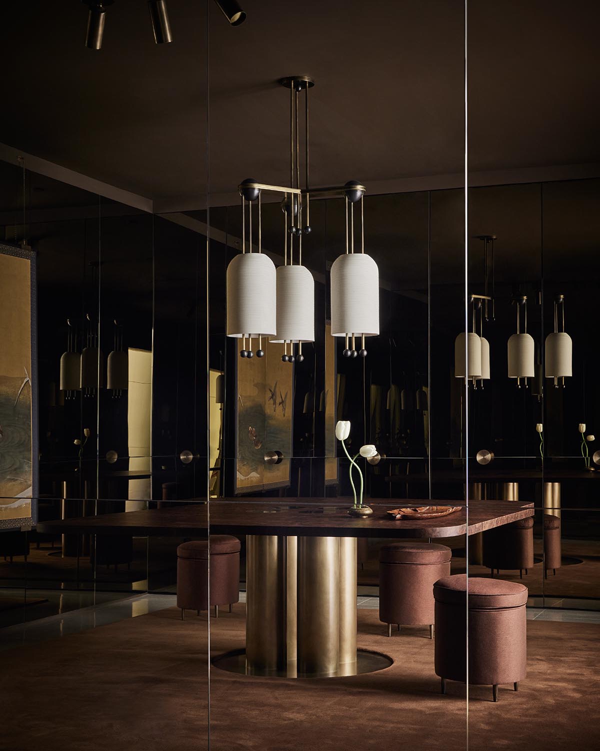 Dark elegant dining space with a wooden table featuring a cylindrical brass base and three upholstered round stools beneath white pendant lights