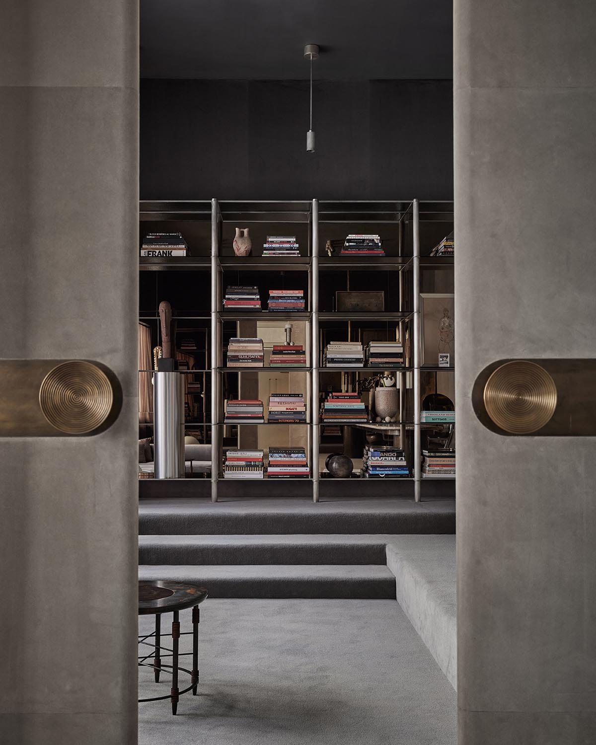 View through open concrete doors with large brass handles into a dim library with a metal bookshelf filled with books and decor objects