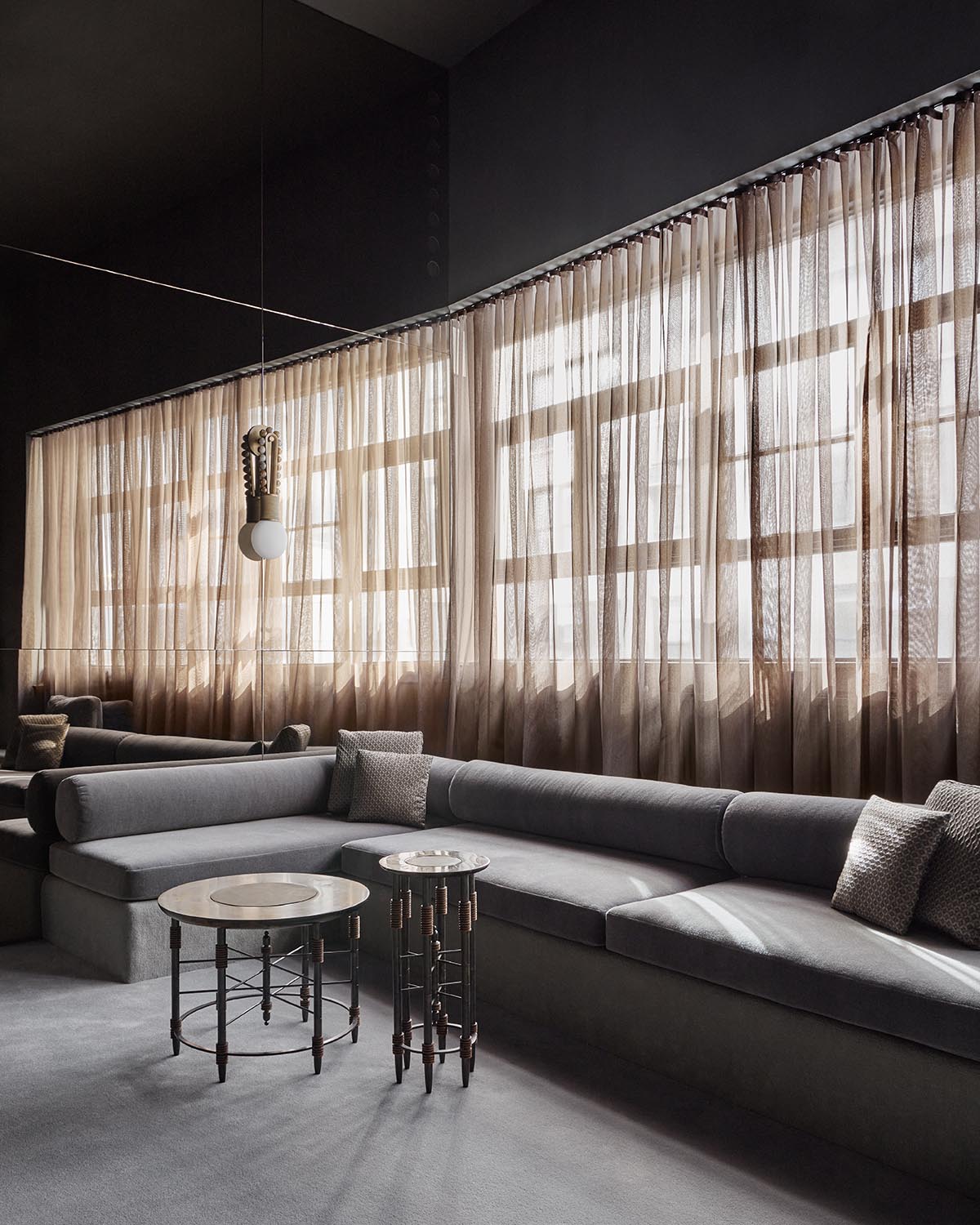 Contemporary lounge area with a long gray sofa, sheer beige curtains, and two round metal side tables on a gray floor