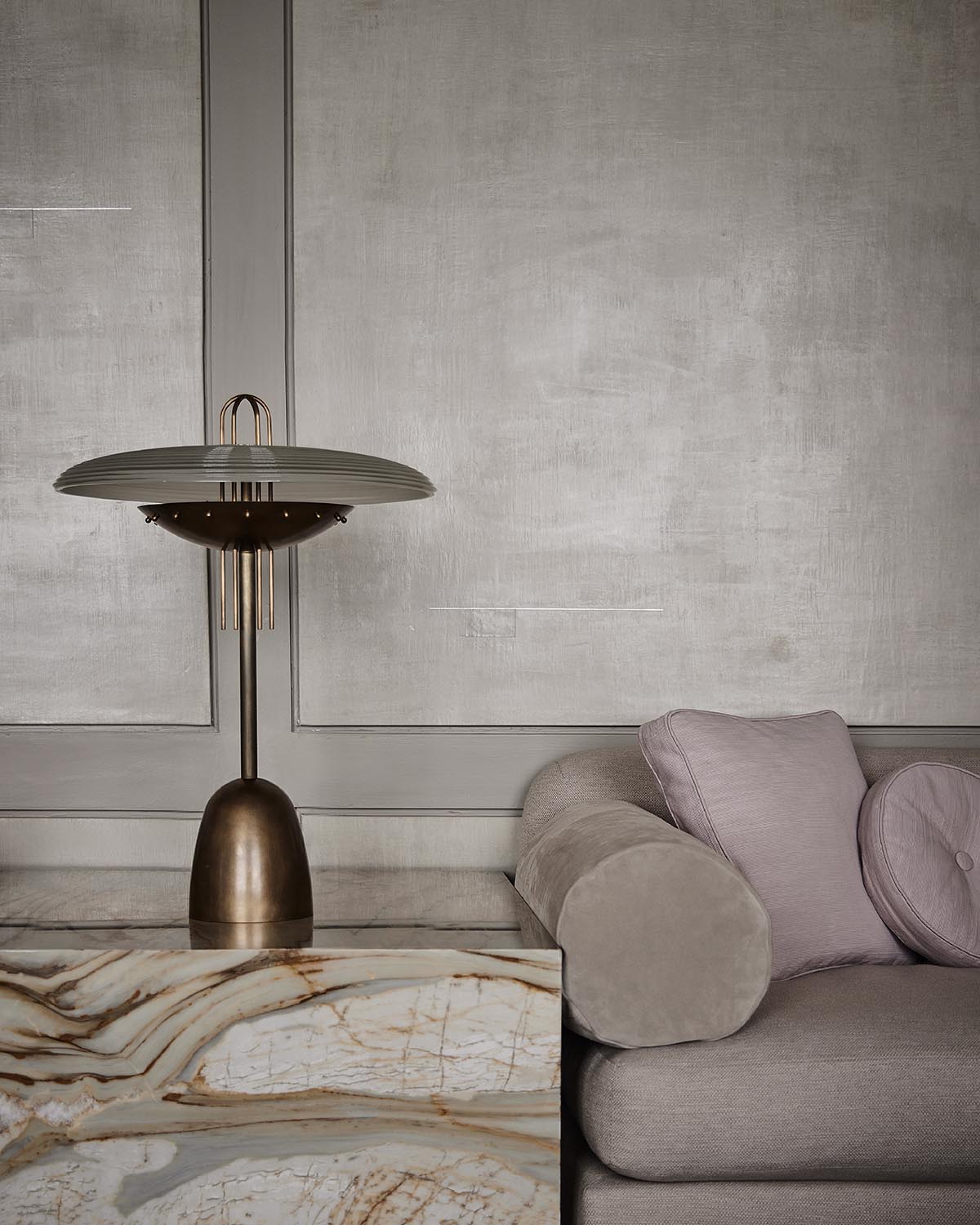 Brass and glass table lamp on a polished marble side table beside a taupe sofa with textured cushions against plastered wall