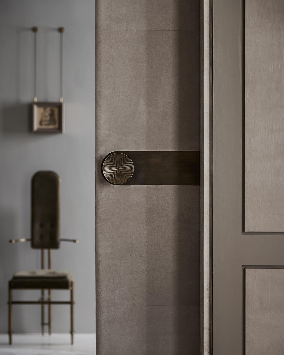 Close-up of a round textured metal door handle on a matte gray door with a modern chair and framed artwork blurred in background