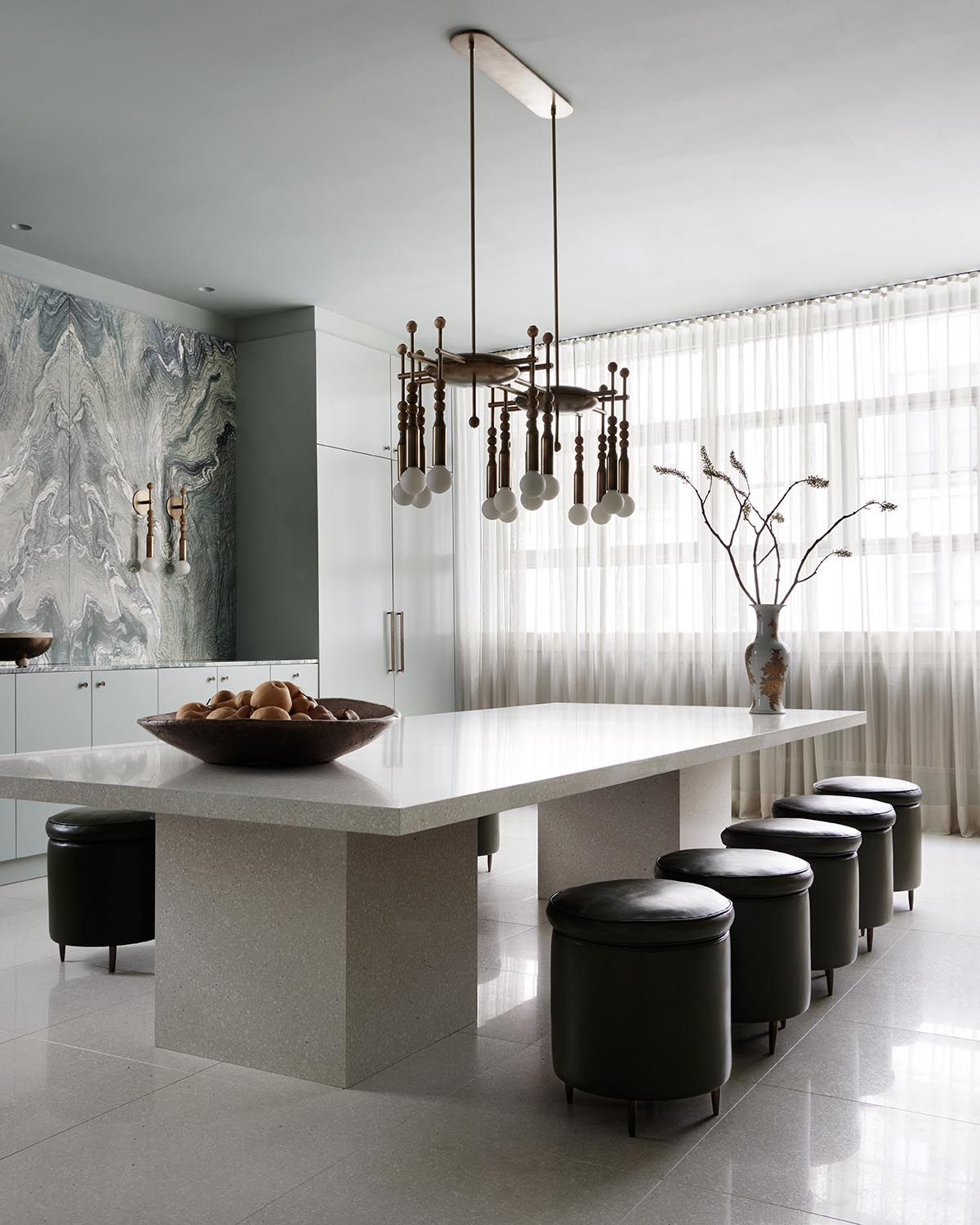 Modern dining area with a large white stone table, black leather stools, marble accent wall, and sheer window curtains
