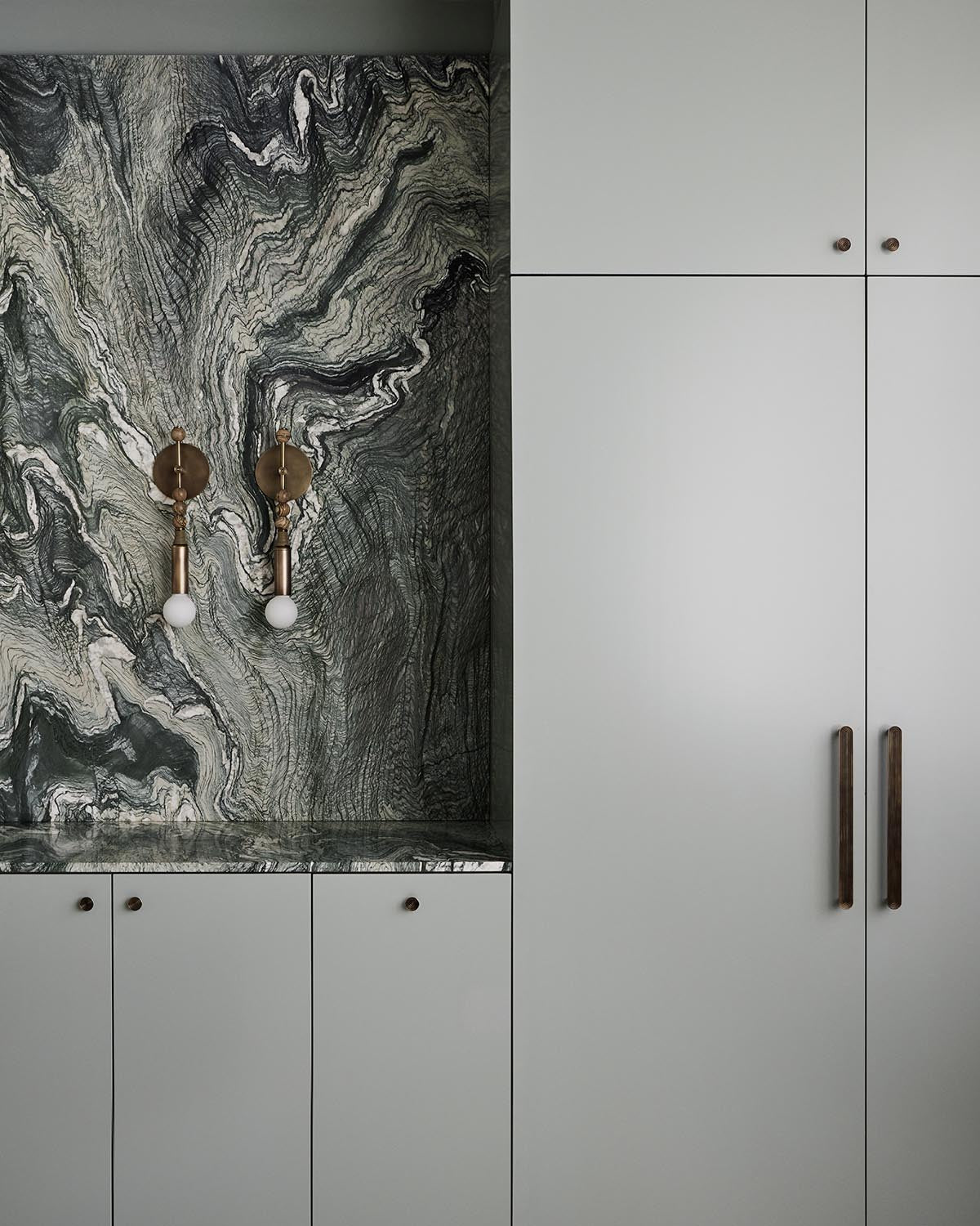 Minimalist kitchen area with light gray cabinetry, dark brass handles, and a dark marble backsplash with swirling patterns.