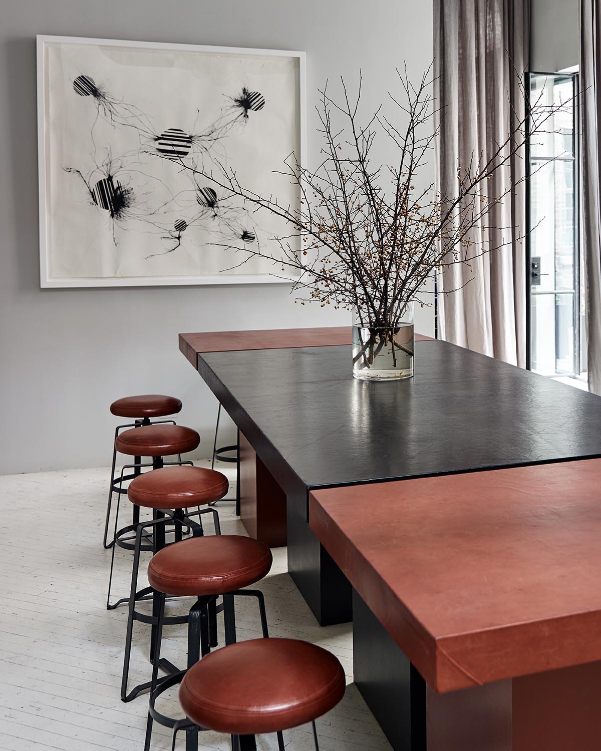 Modern dining area with black and reddish-brown table, round stools, large abstract art, and vase with bare branches
