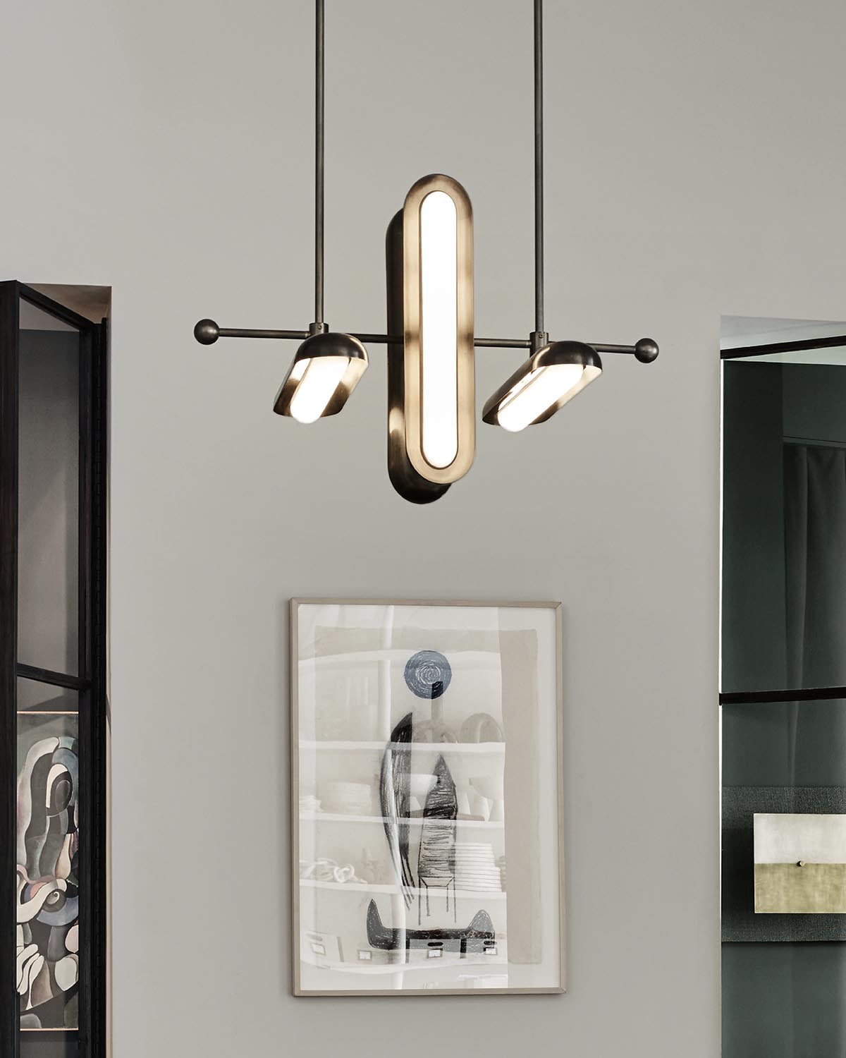 Modern pendant light fixture with elongated oval and round illuminated elements against a light gray wall with framed artwork
