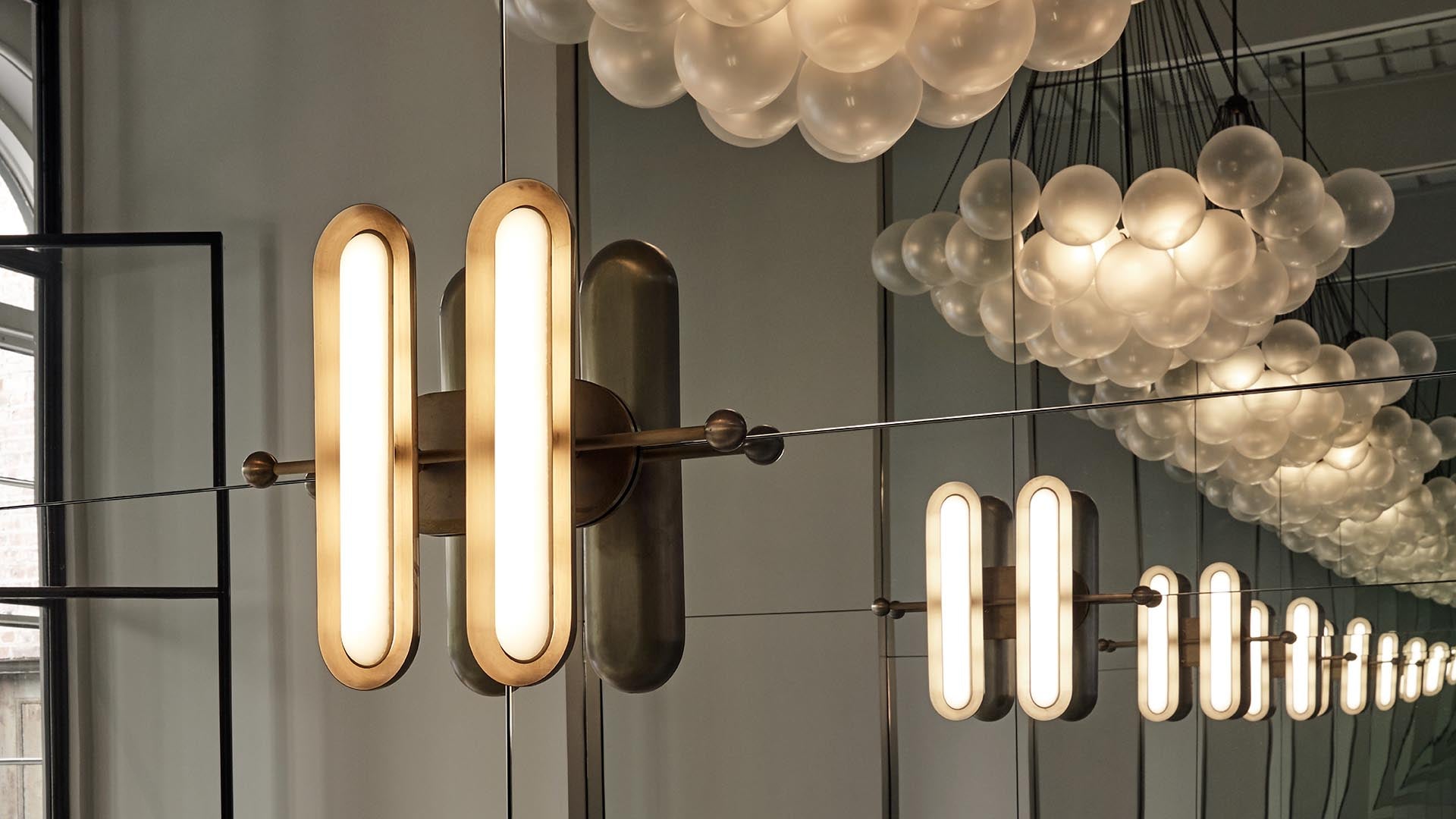 Modern wall light fixture with elongated frosted glass bulbs reflected in a large mirror against a gray wall
