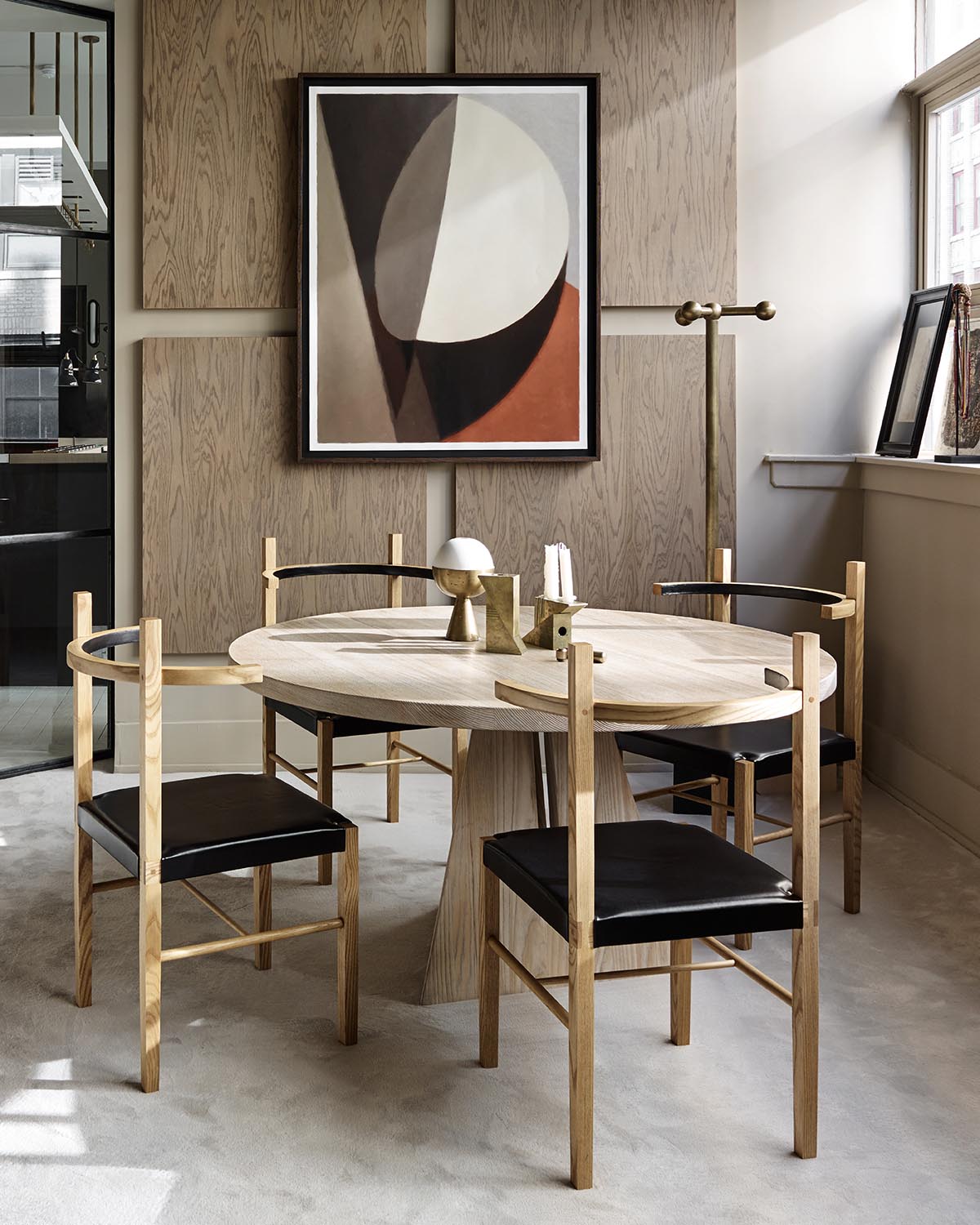 Dining area with round wooden table and four chairs with black seats, abstract wall art, and brass coat rack by a window