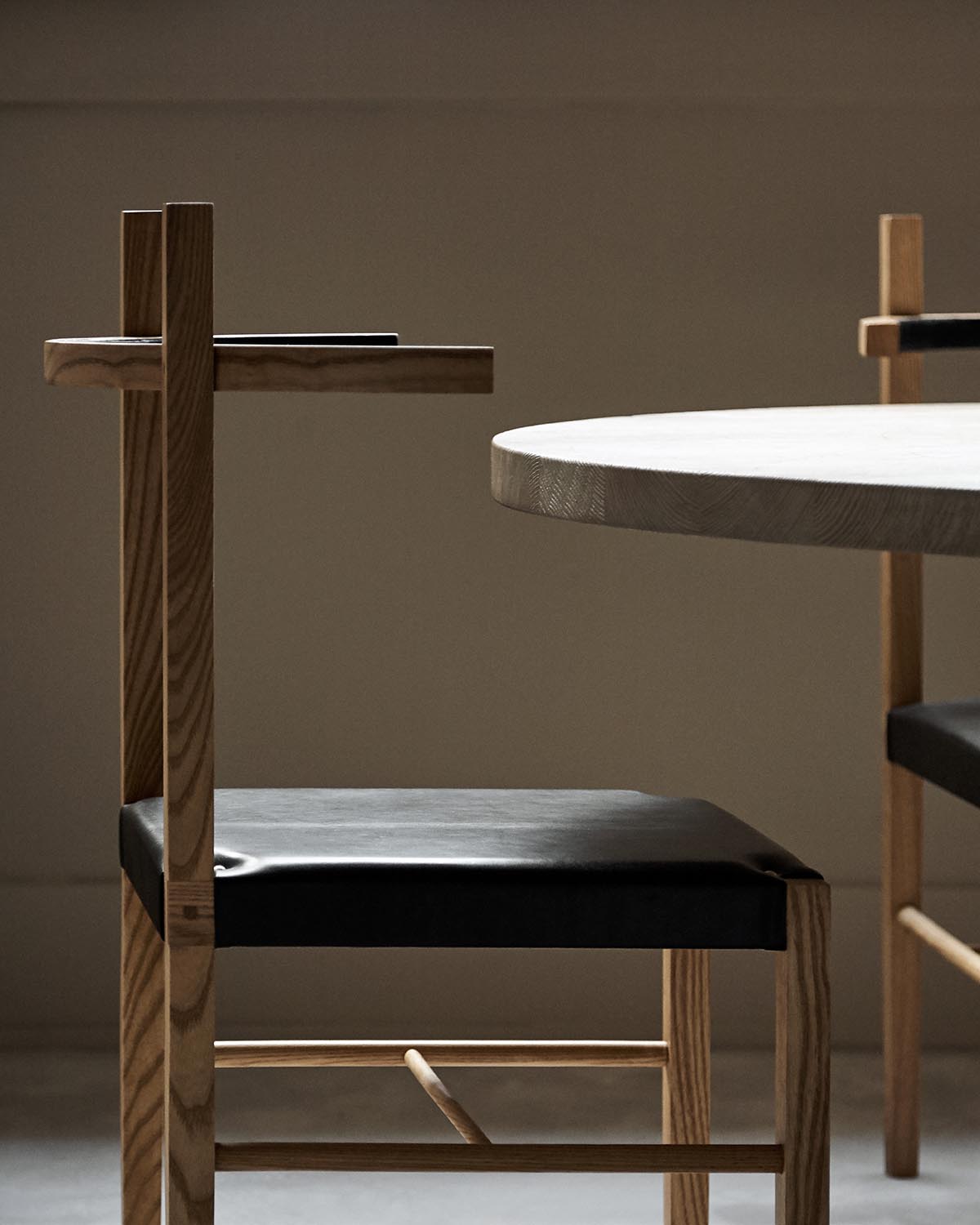 Side view of a wooden chair with black leather seat next to a round light wood table against a muted background
