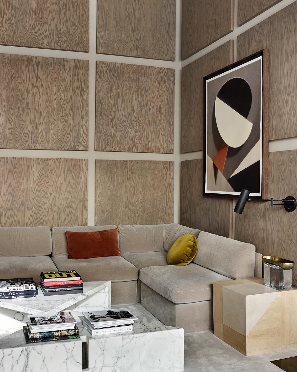 Contemporary living room with beige sectional sofa, geometric marble coffee table, abstract wall art, and wood-paneled walls.