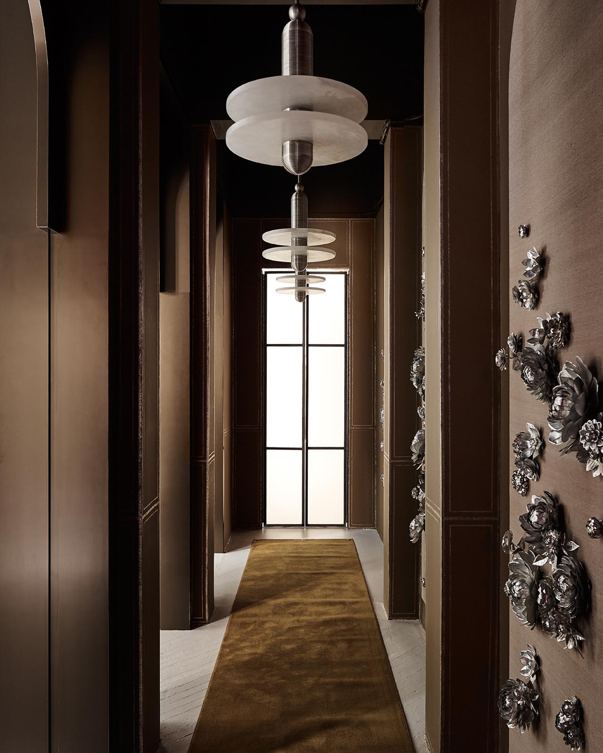 Narrow hallway with brown walls, metallic floral wall sculptures, pendant lights, and a window with frosted glass panels