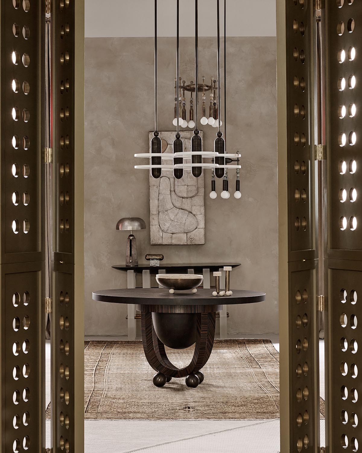 Interior view through ornate perforated doors revealing a round wooden table with sculptural base on a woven rug beneath modern pendant lights