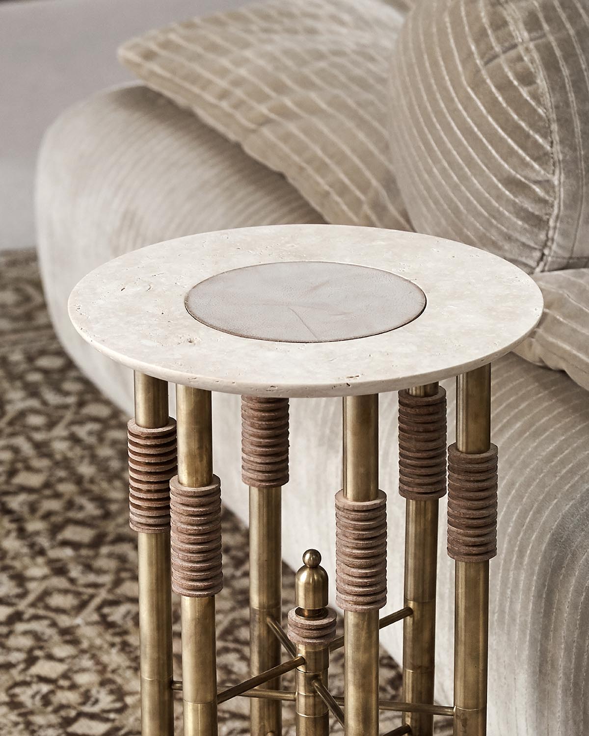 Close-up of a round side table with a light stone top and brass legs wrapped in textured brown rings beside a beige corduroy sofa
