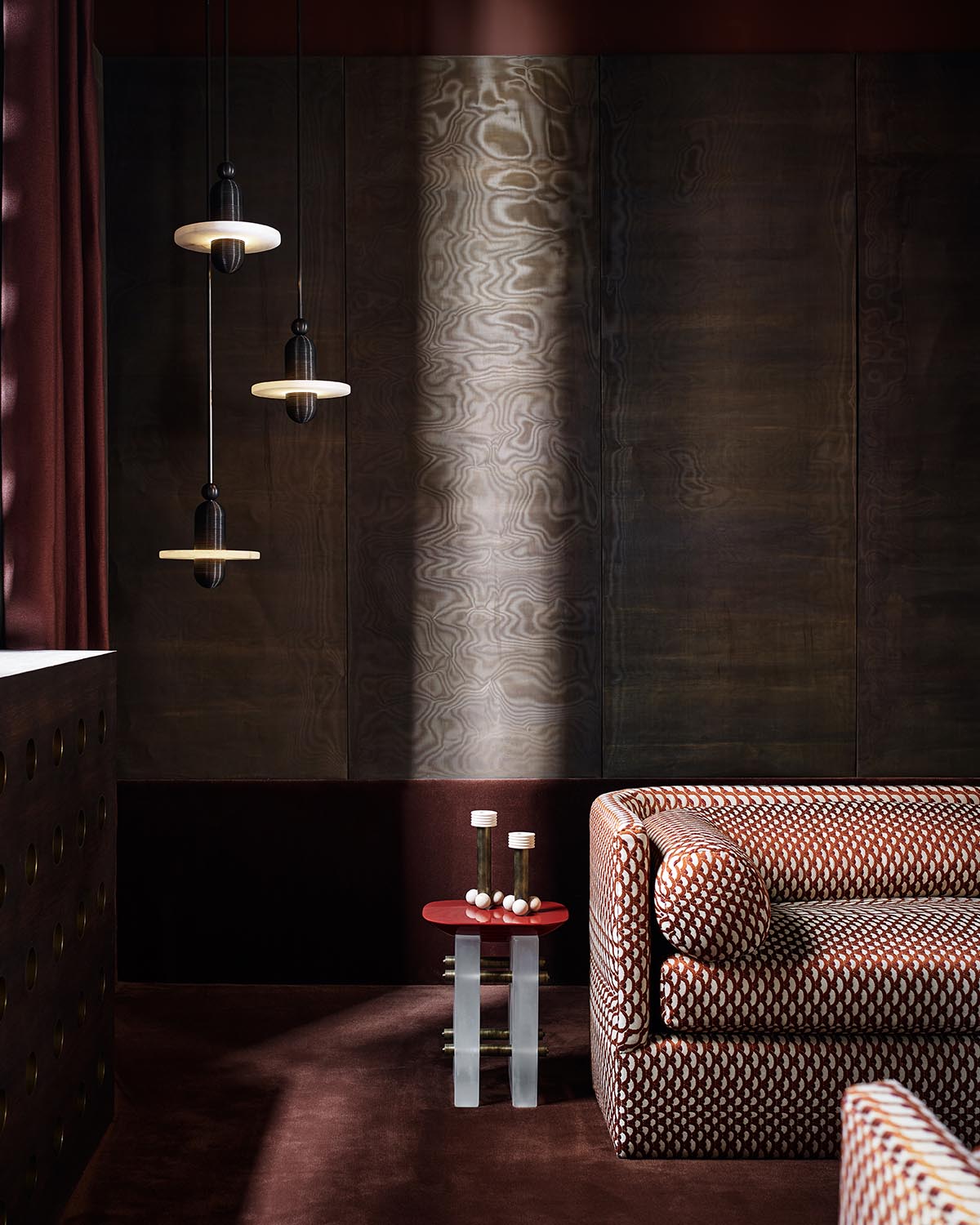 Corner of a room with patterned upholstered sofa, small red side table holding candles, and three hanging pendant lights against dark wood walls