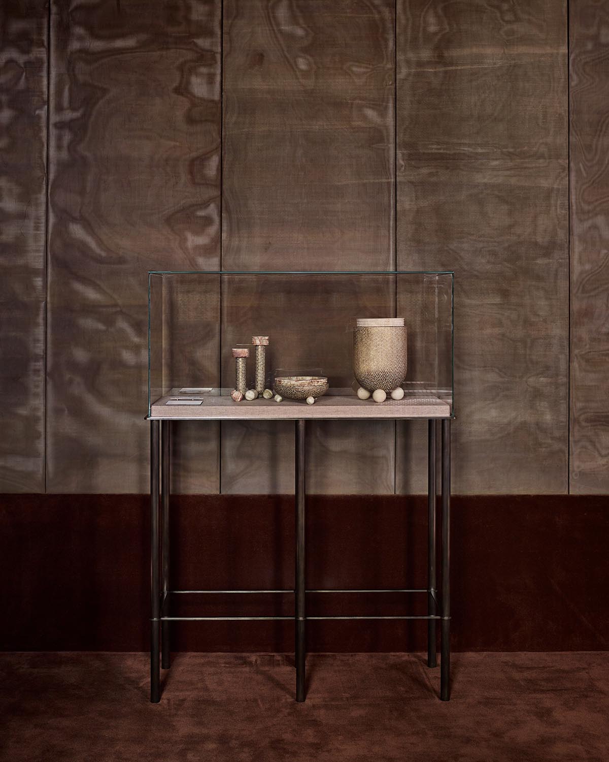 Glass display case on a slender metal table showcasing textured cylindrical and round objects against a wood-paneled wall