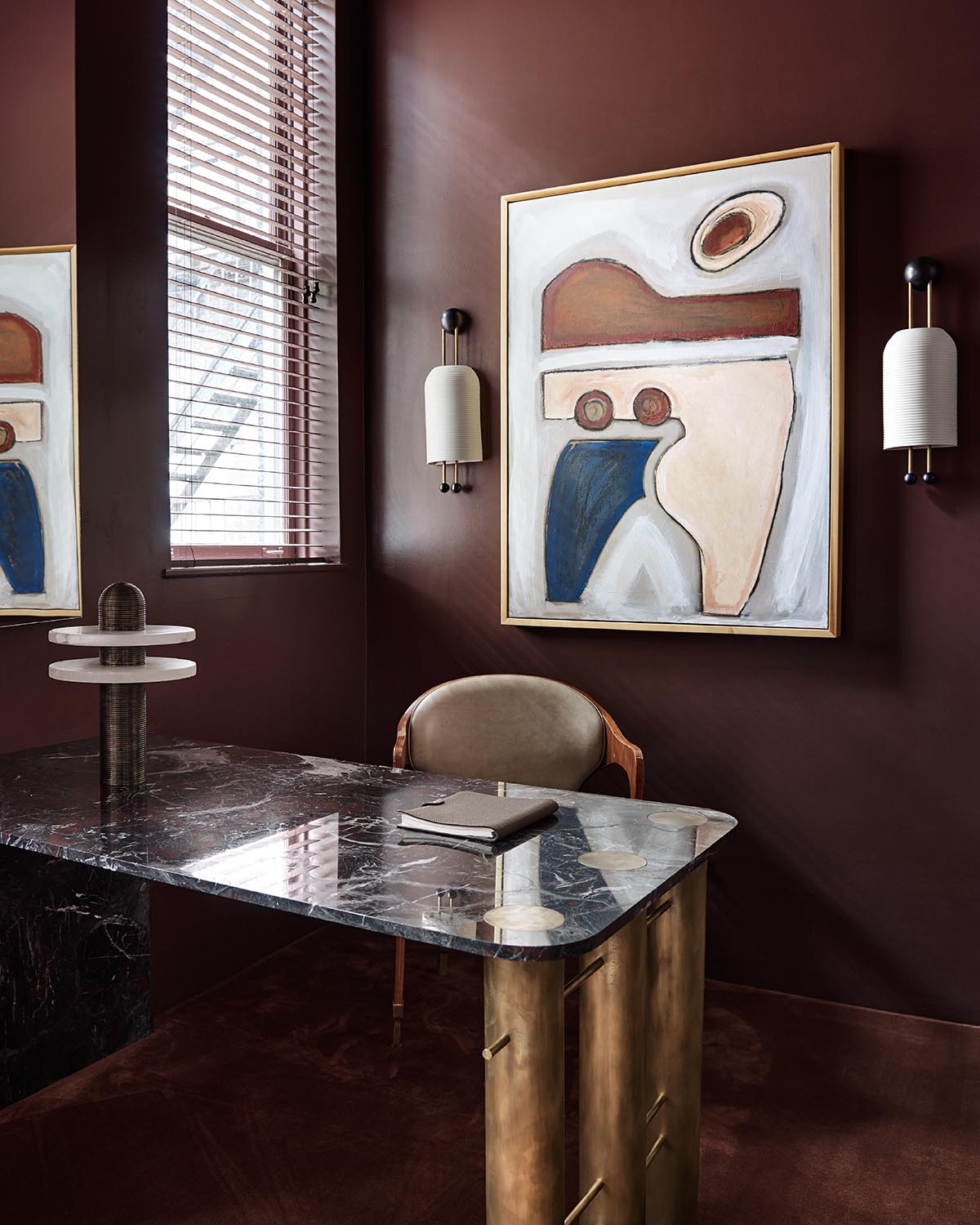 Modern office corner with dark marble desk, beige chair, abstract painting, and frosted wall sconces on brown walls
