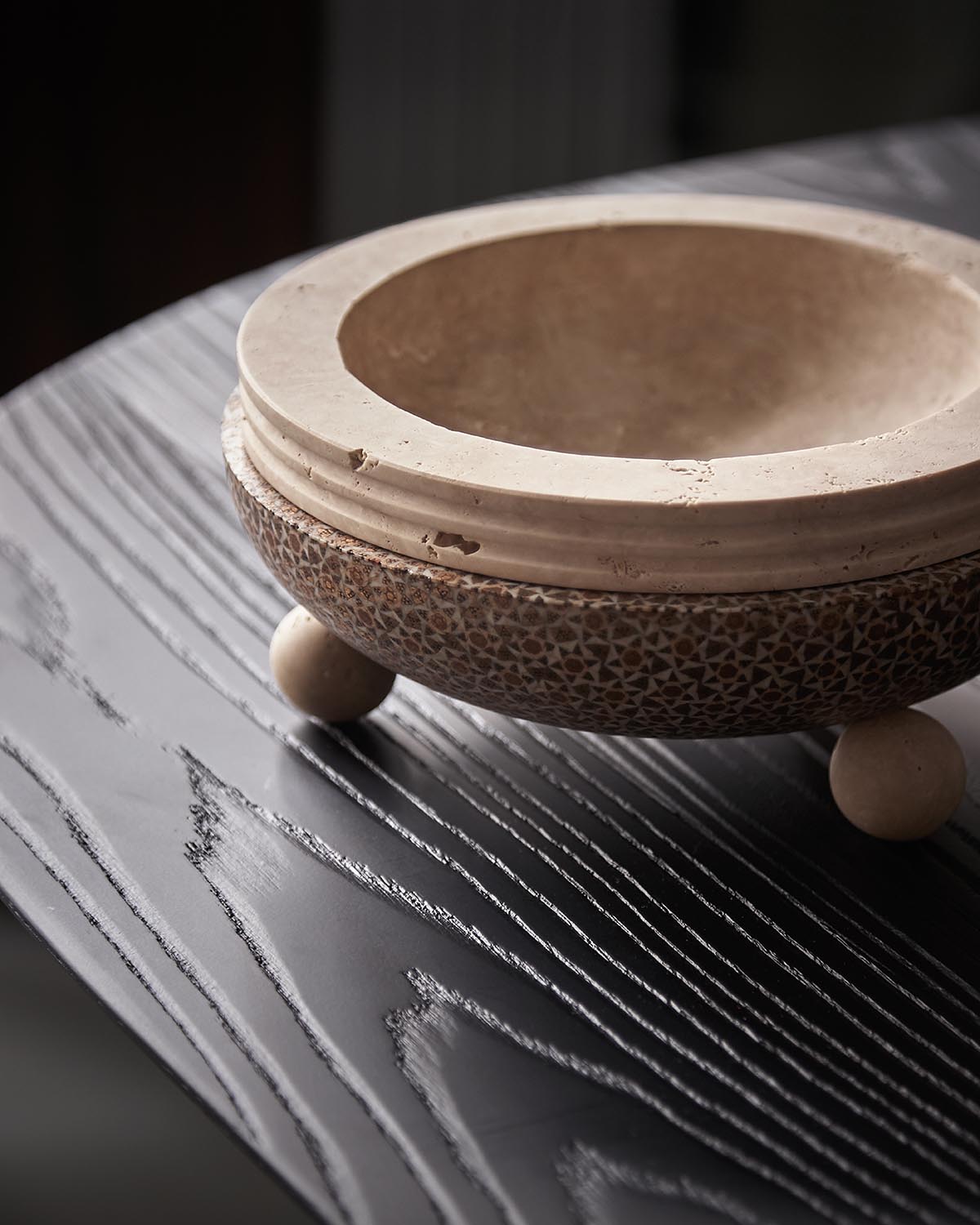 Close-up of a round textured stone bowl with small ball feet placed on a dark wood surface with visible grain patterns