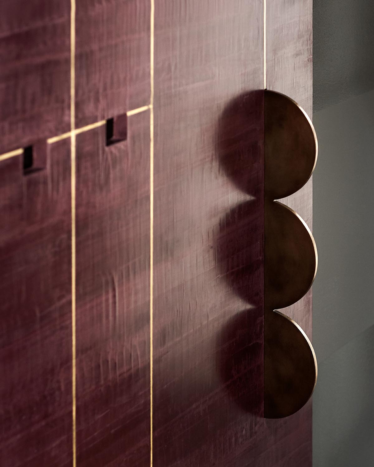 Close-up of dark wood paneling with vertical gold inlays and three round metallic handles casting shadows on a gray wall background