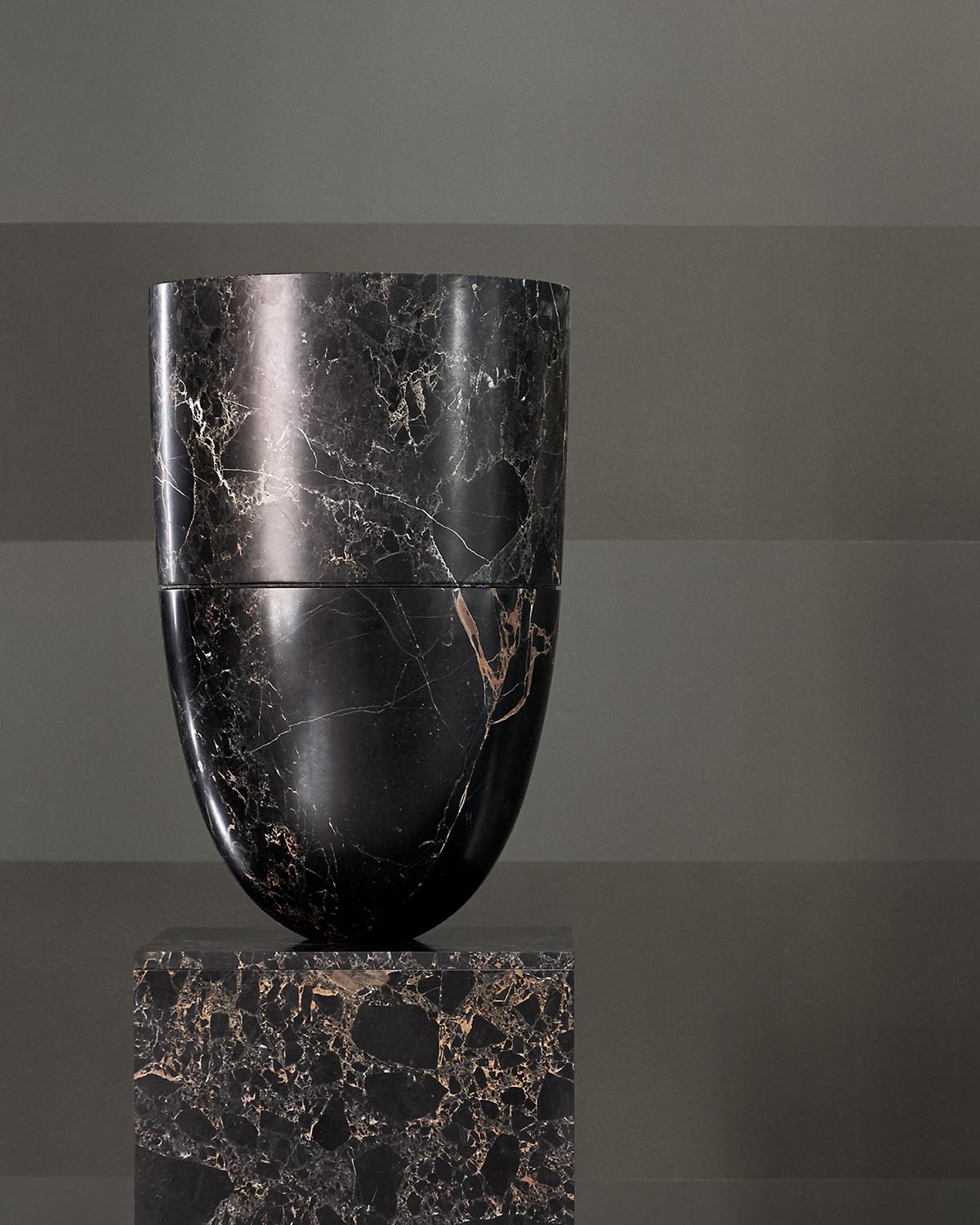 Tall black marble vase with beige and white veins standing on matching square marble pedestal against a gray background