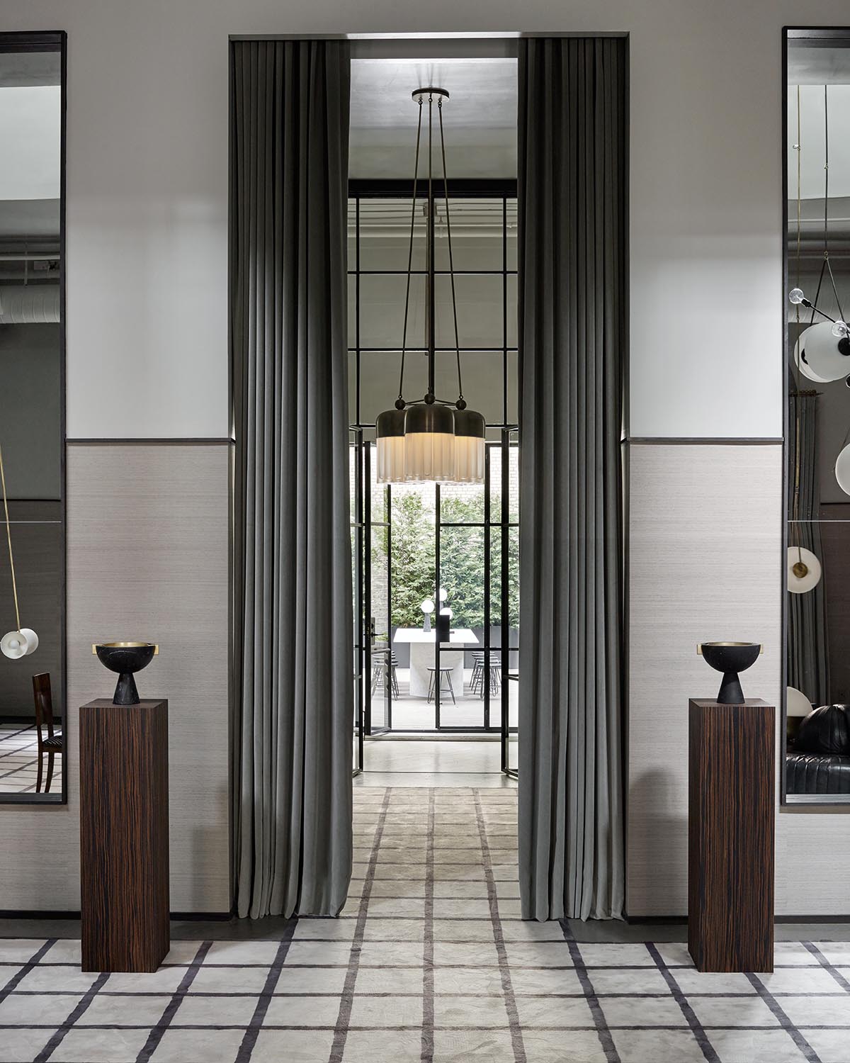 Symmetrical modern interior with tall gray curtains, dark wood pedestals holding black bowls, and a hanging chandelier.
