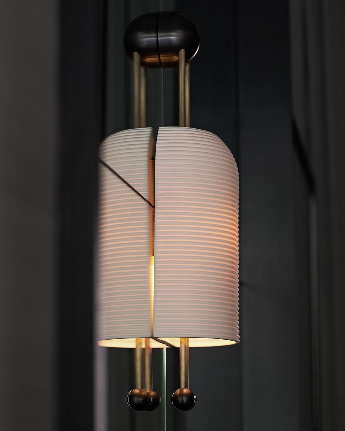 Close-up of a modern pendant lamp with a ribbed white cylindrical shade and brass vertical rods against a dark background