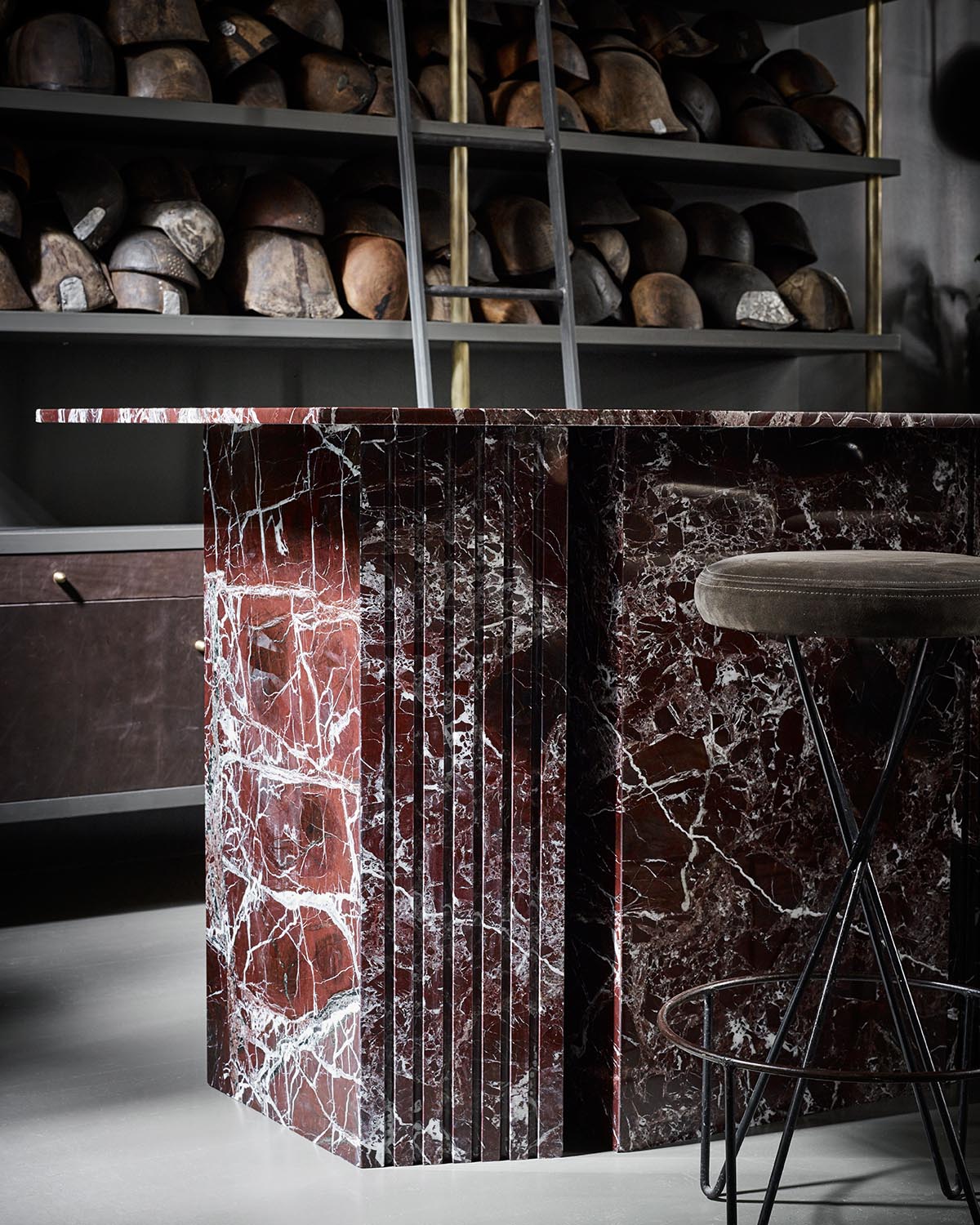 Close-up of a dark red marble bar with white veining, accompanied by a cushioned stool with metal legs and wooden shelves in the background