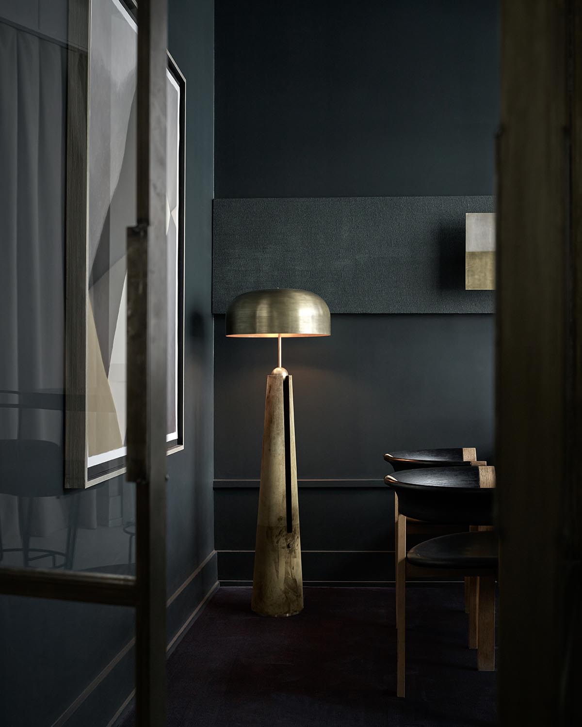 Tall brass floor lamp with rounded shade standing beside dark wooden chairs in a dimly lit modern interior room