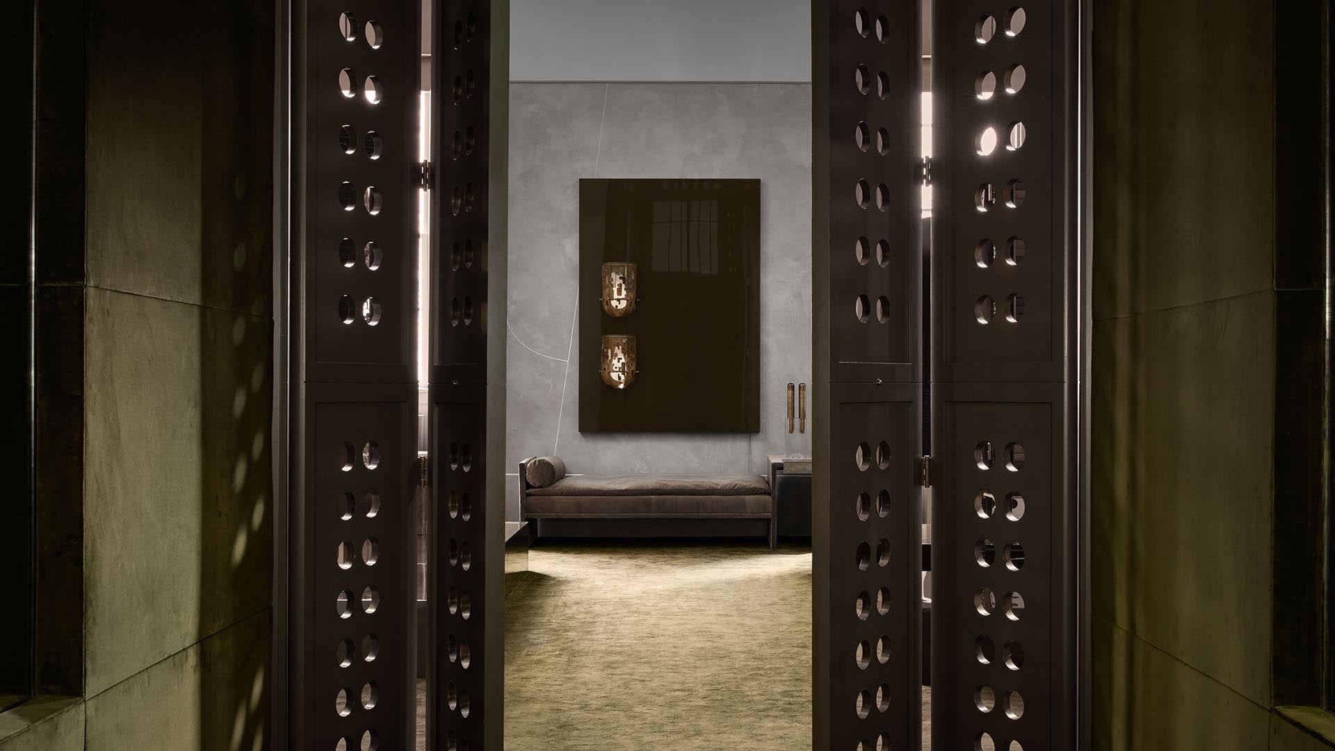 View through perforated dark metal panels into a minimalist room with a bench, textured wall, and modern lighting fixtures