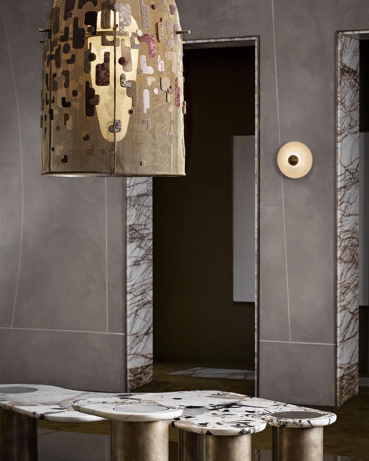 Abstract pendant light with gold and brown mosaic pattern hanging above round marble tables with cylindrical bases in a muted interior