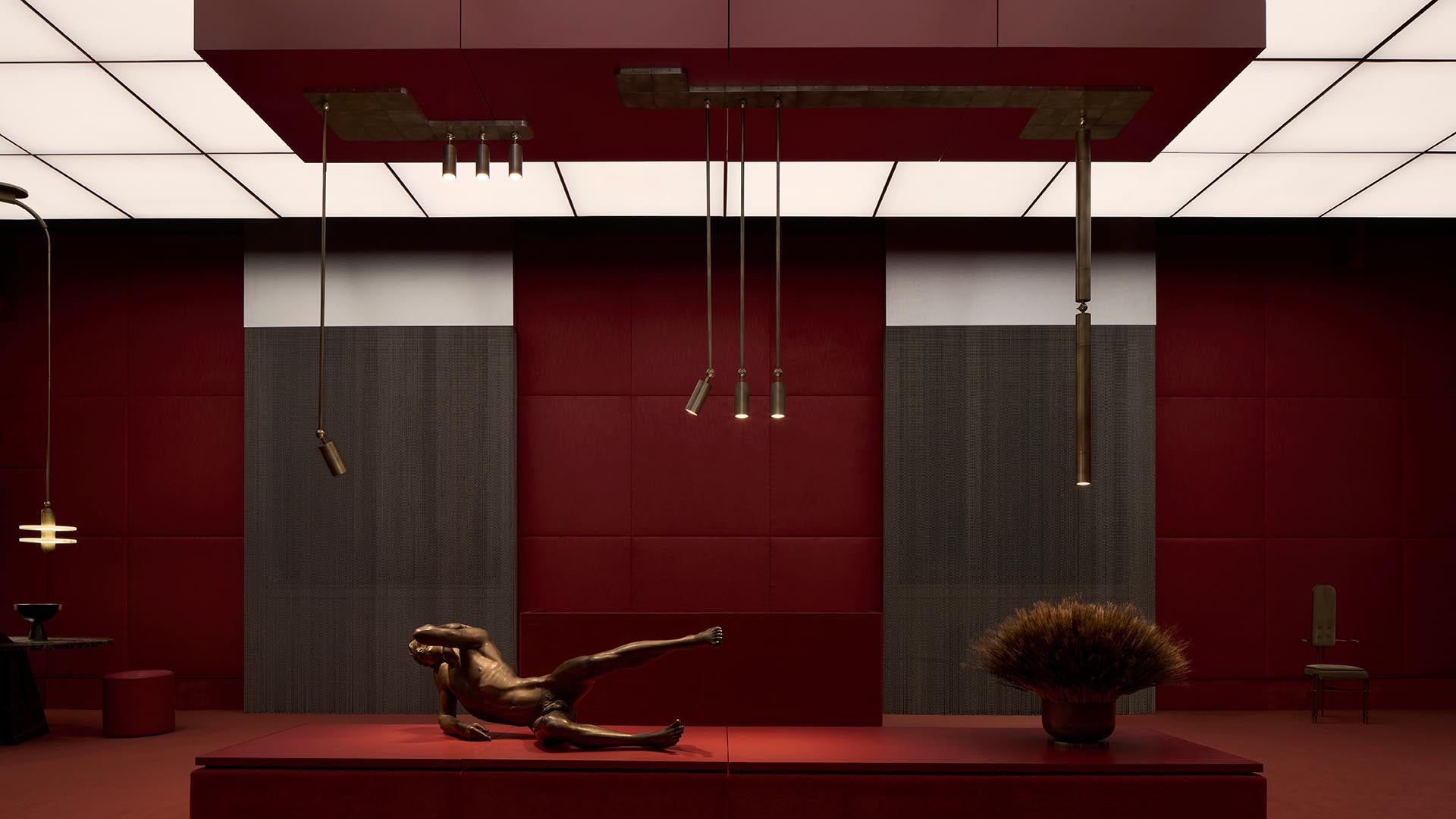 Dimly lit modern interior with dark red walls, ceiling panels, hanging brass fixtures, and a reclining bronze sculpture on a platform