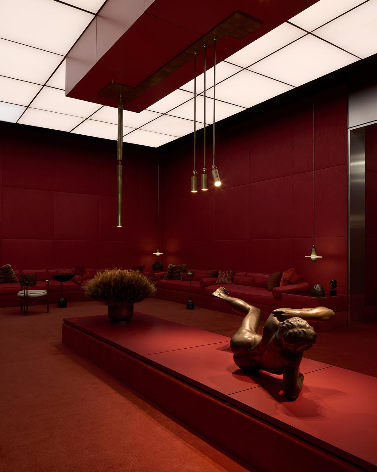Dimly lit room with deep red walls, a bronze reclining sculpture on a low platform, and modern pendant lights above.