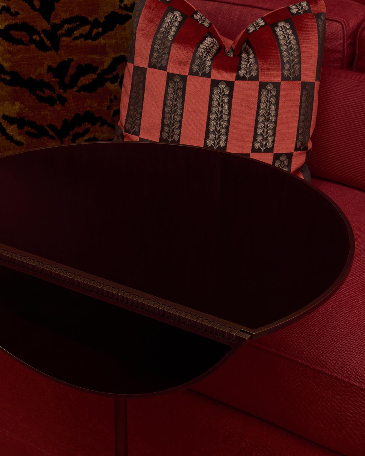 Close-up of a dark oval tabletop with a metallic hinge detail, set against a red fabric sofa and patterned cushion backdrop